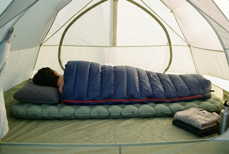 A camper asleep on their side in a tent, seen from behind, on a thick green sleeping pad with a quilted pillow and a navy down sleeping bag.