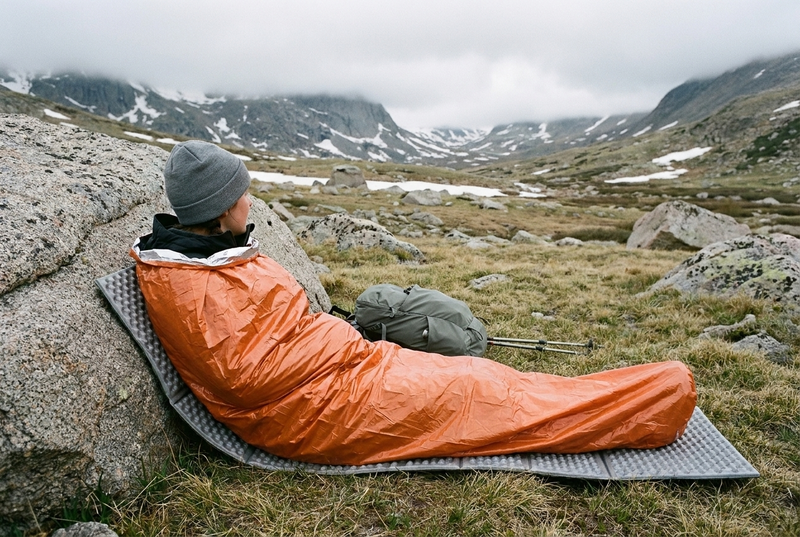 A hiker in a gray beanie, seen from behind, half lying against a granite boulder in a snow-patched alpine valley, in an orange emergency bivvy sack pulled up to the shoulders, on a gray foam pad under the whole body, with a daypack and trekking poles.