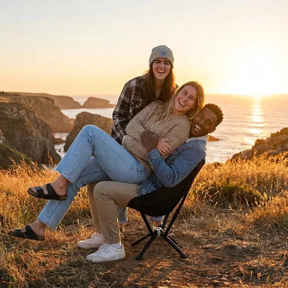 Three friends sharing a Restwild folding camping chair on a cliff at sunset
