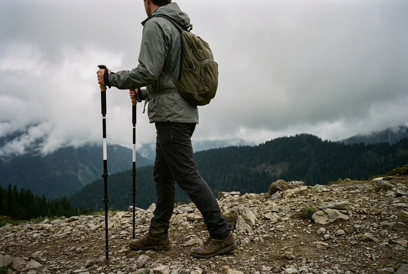 A hiker in a gray-green rain jacket with an olive daypack, seen from behind and to the side on a rocky mountain trail under low cloud, holding a trekking pole with a cork grip in each hand with elbows bent.