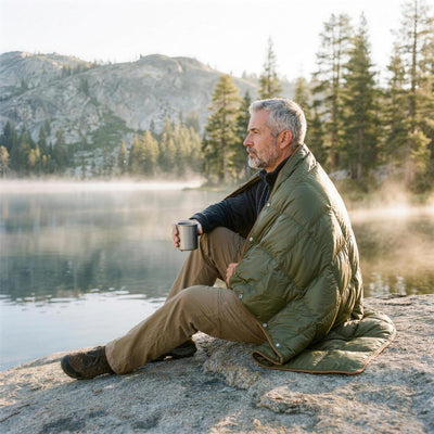 Man sitting on a granite slab by a misty mountain lake with the green puffer blanket wrapped around his shoulders