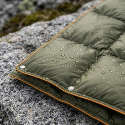 Water drops beading on the green ripstop shell of the puffer blanket, with snap buttons along the orange piped edge
