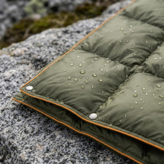 Water drops beading on the green ripstop shell of the puffer blanket, with snap buttons along the orange piped edge