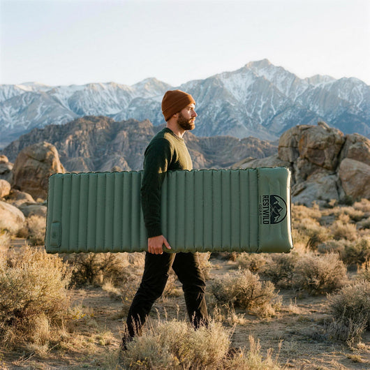 Man carrying an inflated green Mattress Pro V2 sleeping pad under his arm across a sagebrush flat below a snowy mountain range