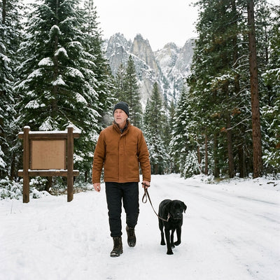 Man in black fleece-lined winter pants walking a black Labrador on a snowy forest road