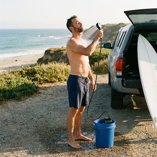 Man rinsing off with the camp shower beside a pickup and a surfboard at the coast, the pump standing in a bucket