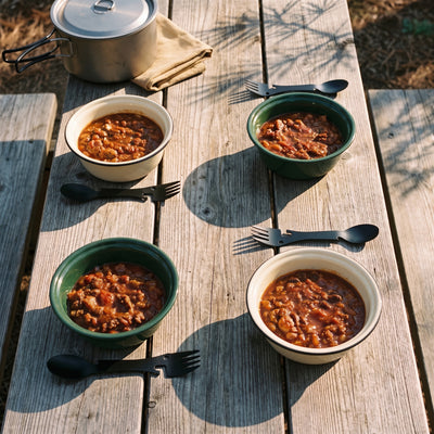 Four bowls of chili on a campground picnic table, each with its own black camping spork