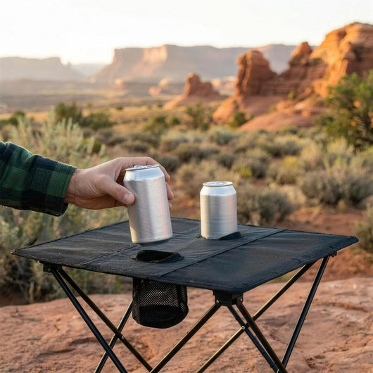 Hand lifting a can out of one of the two cup holders on the folding camping table, desert mesas behind