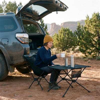 Woman in a camp chair beside an SUV at a desert trailhead, taking a mug from the folding camping table